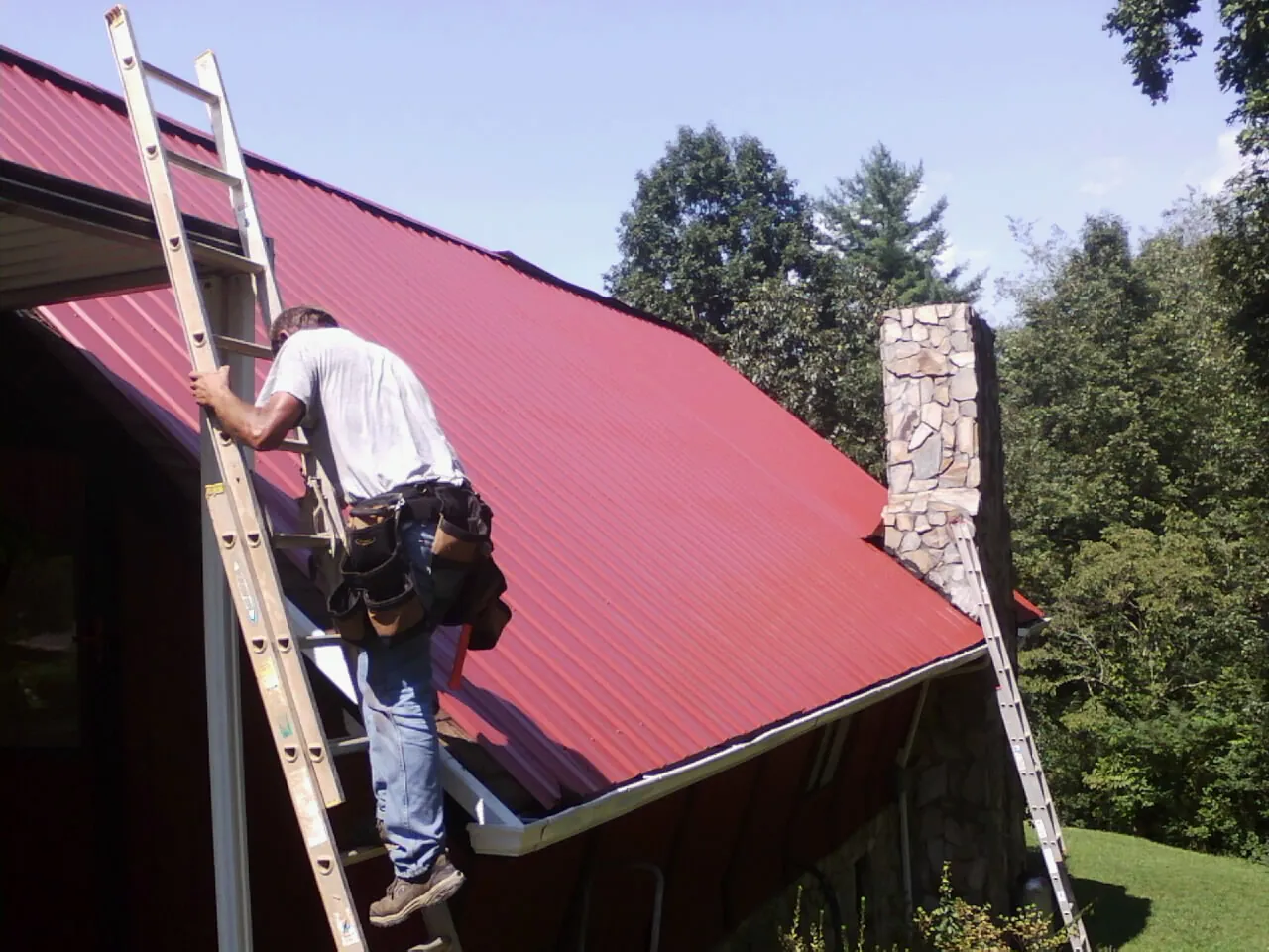 Roofer on a ladder beside a red metal roof and stone chimney — Soffit & Fascia Repair by Roofing Brothers in Warwick